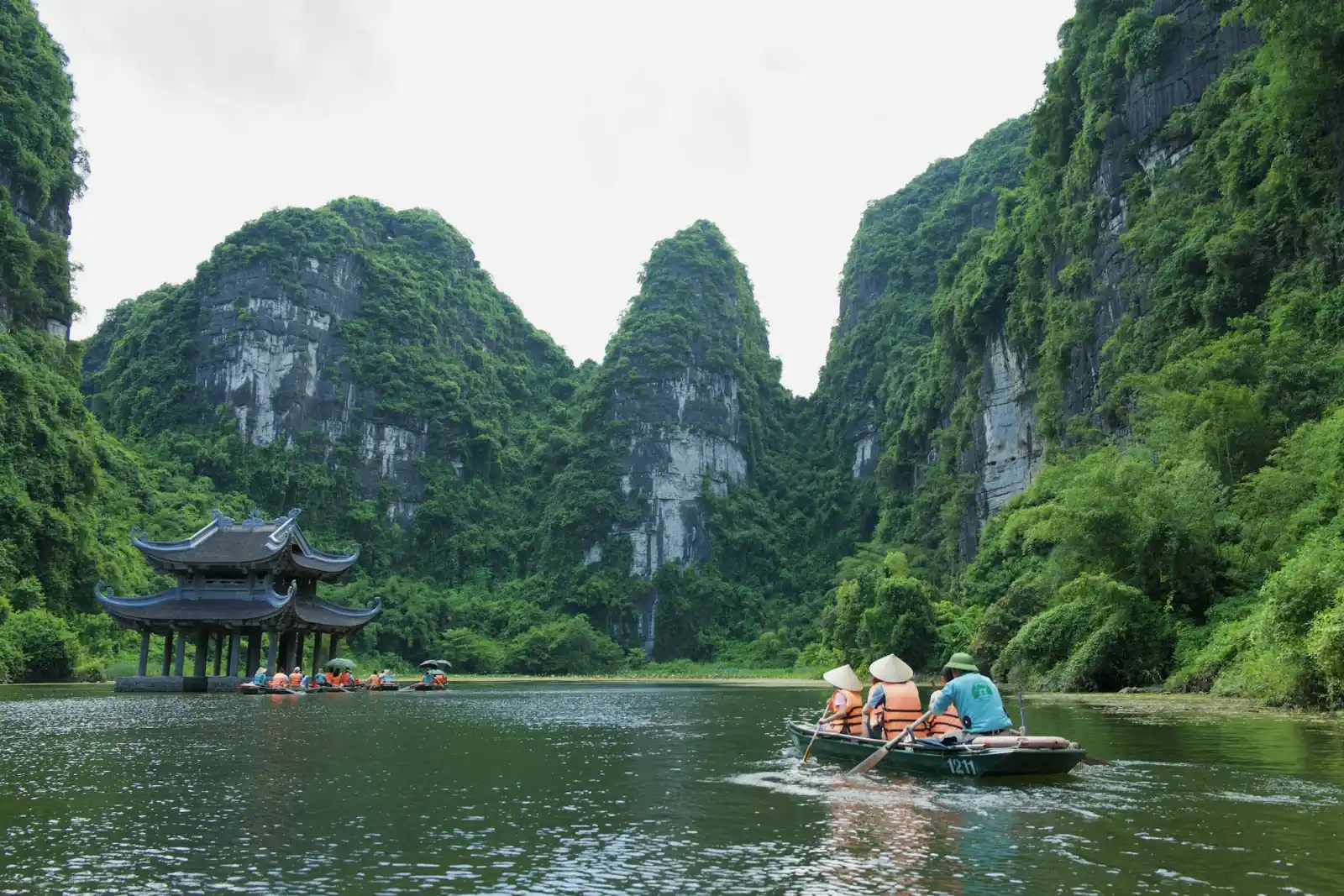 Thuyền chở khách trên sông giữa những dãy núi đá vôi, bên bờ là ngôi đình nhỏ